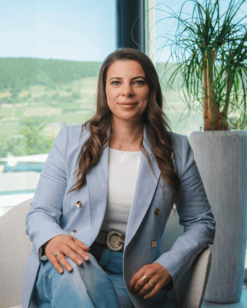 Porträt einer jungen Frau im hellblauen Blazer, sitzend vor Fenster mit Ausblick ins Grüne – Ansprechpartnerin für das Neubauprojekt Rheintal47