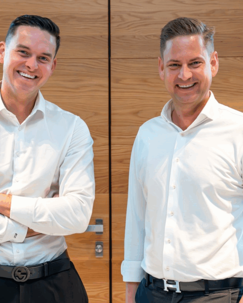 Zwei freundlich lächelnde Männer in weißem Hemd vor einer Holzwand – Ansprechpartner für das Neubauprojekt Rheintal47 in Bingen